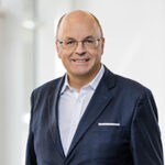 Portrait de Stephan Hofstetter en costume avec lunettes, souriant sur un fond neutre.