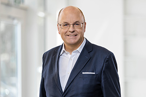Porträt von Stephan Hofstetter in einem Anzug mit Brille, lächelnd vor neutralem Hintergrund.