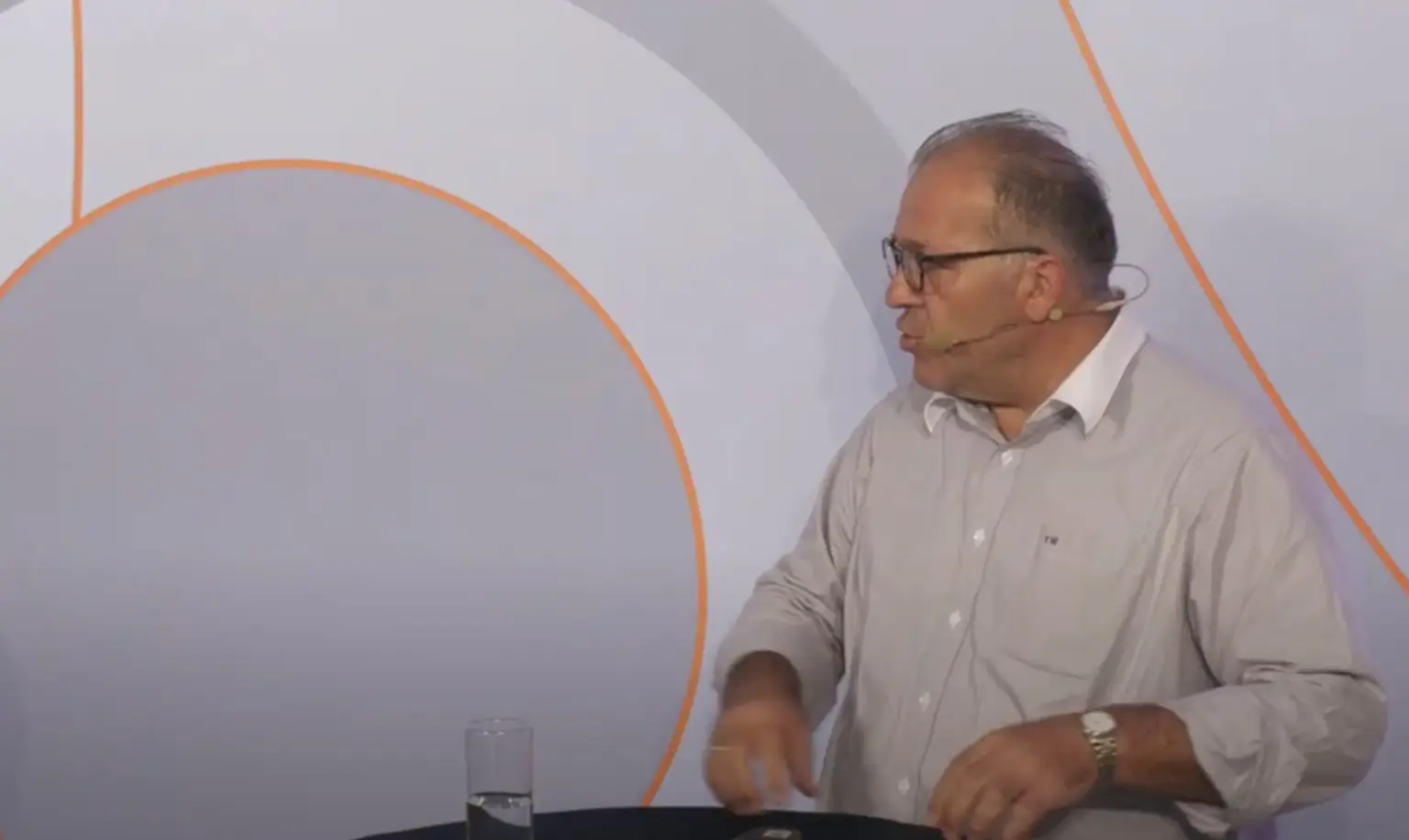 Mann mit Brille und Mikrofon spricht am Tisch mit einem Glas Wasser.