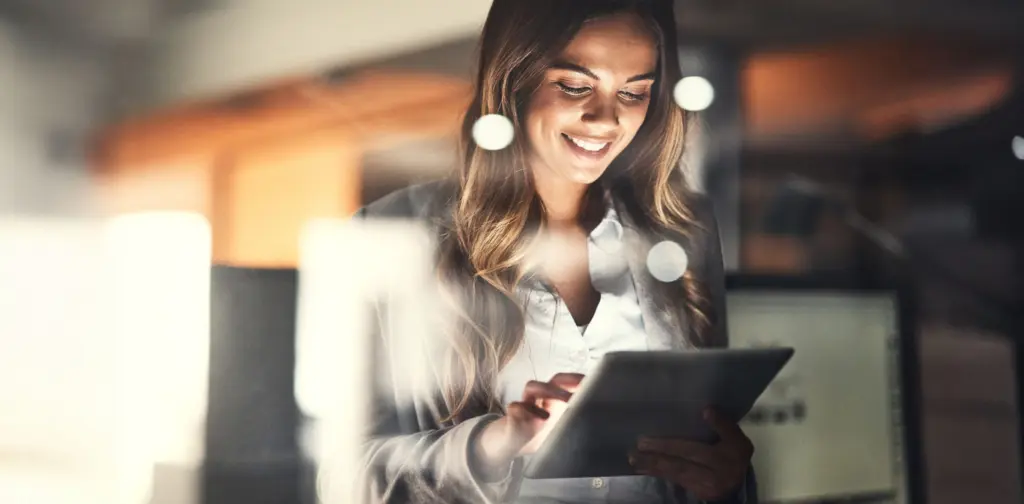 Une femme sourit en tenant une tablette dans un bureau et en tapant dessus.
