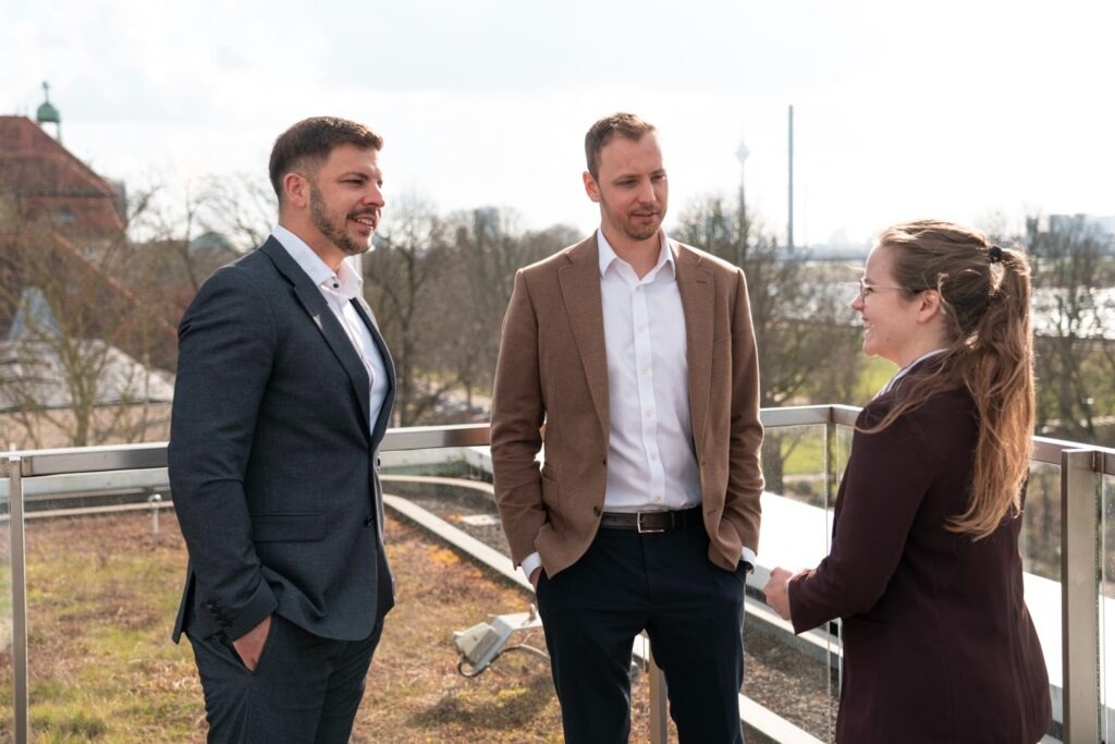 Drei Personen, zwei Männer und eine Frau, stehen auf einer Terrasse und unterhalten sich.
