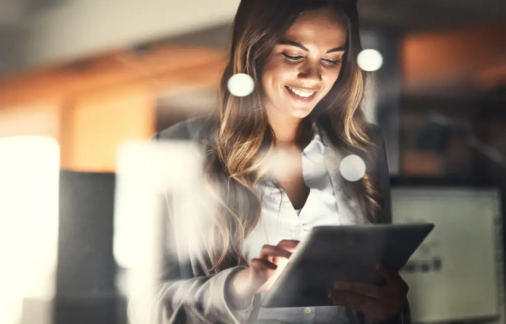 Frau mit langen Haaren, die in einem modernen Büro ein Tablet benutzt und lächelt.