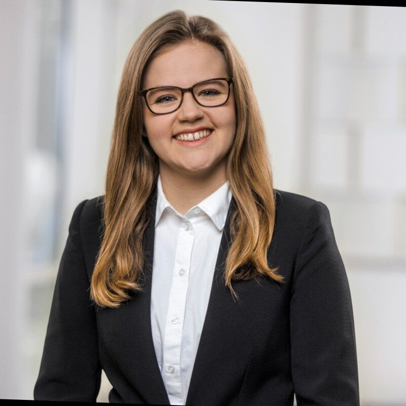 Eine Person mit langen Haaren und Brille, die in einem formellen Outfit lächelt.
