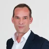 Porträt eines Mannes in einem dunklen Anzug mit offenem Hemdkragen vor einem neutralen Hintergrund.
