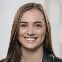 Portrait einer Frau mit langen, braunen Haaren und einem Lächeln, die in einem schwarzen Blazer gekleidet ist.