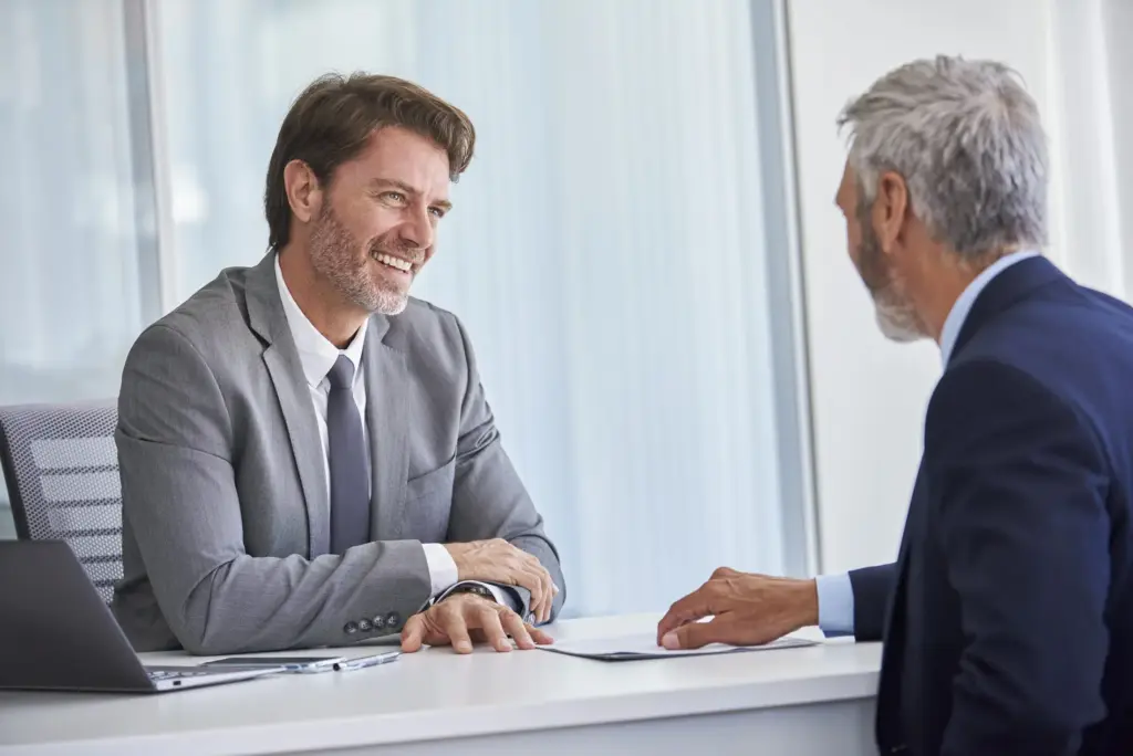 Two men in formal clothing are sitting at a desk having a discussion.