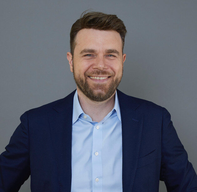 Portrait d'un homme barbu, souriant, vêtu d'un costume et d'une chemise.