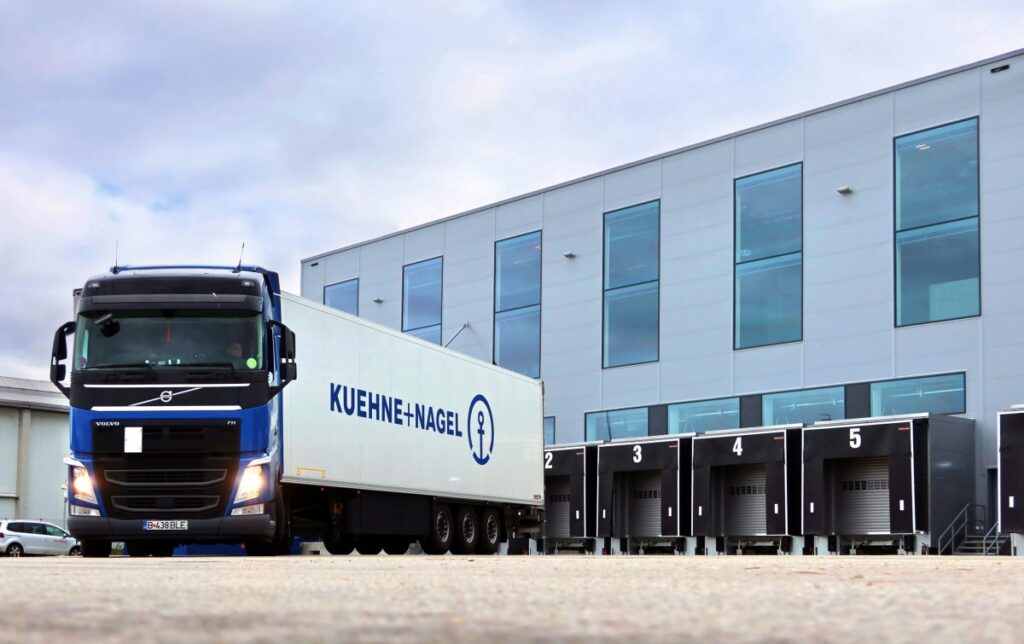 Ein blauer Lkw von Kuehne + Nagel steht vor einem Lagergebäude mit mehreren Ladebuchten.