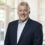 A man in a suit with grey hair and a friendly smile stands in front of a bright wall.