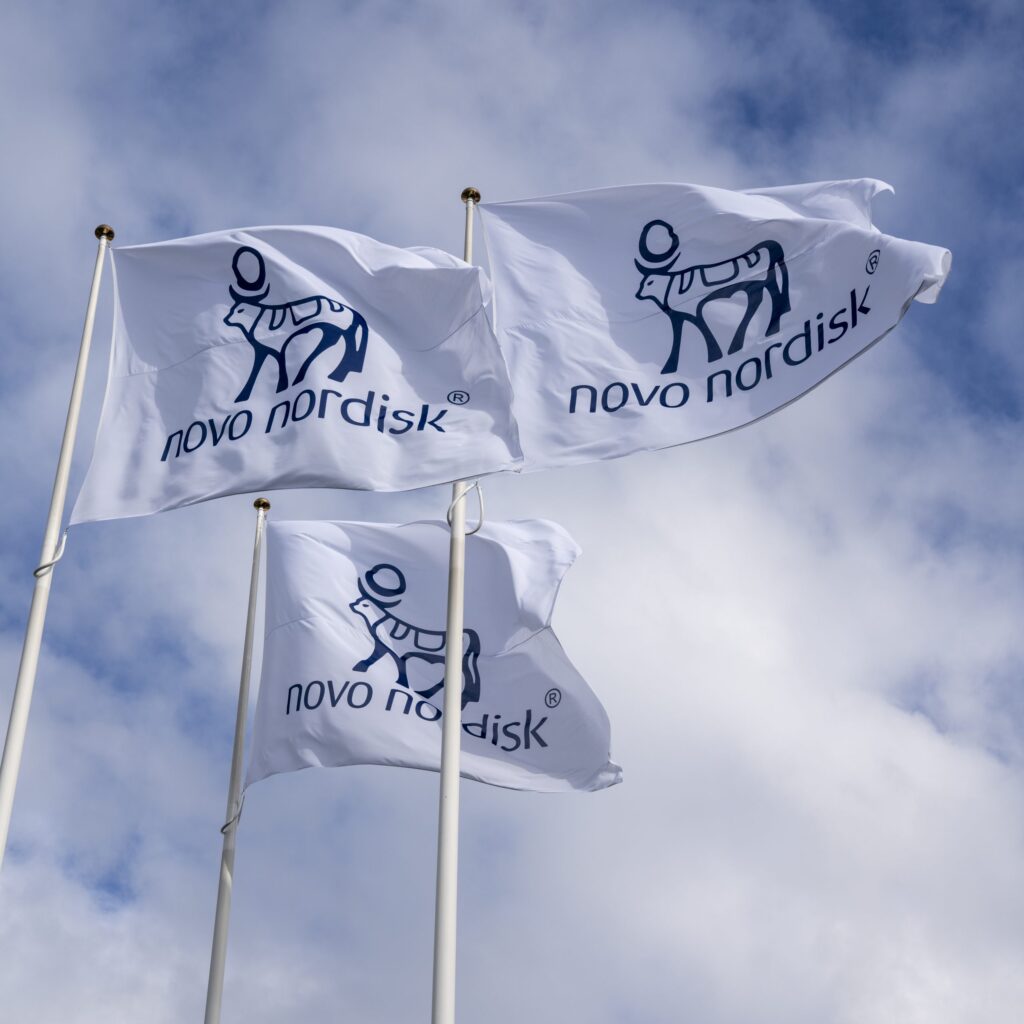 Trois drapeaux de Novo Nordisk flottant au vent, avec le logo sur fond blanc.