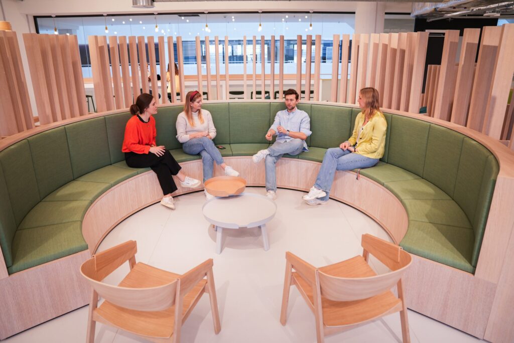 Vier Personen sitzen in einem modernen Besprechungsraum auf einer runden, gepolsterten Sitzgelegenheit.