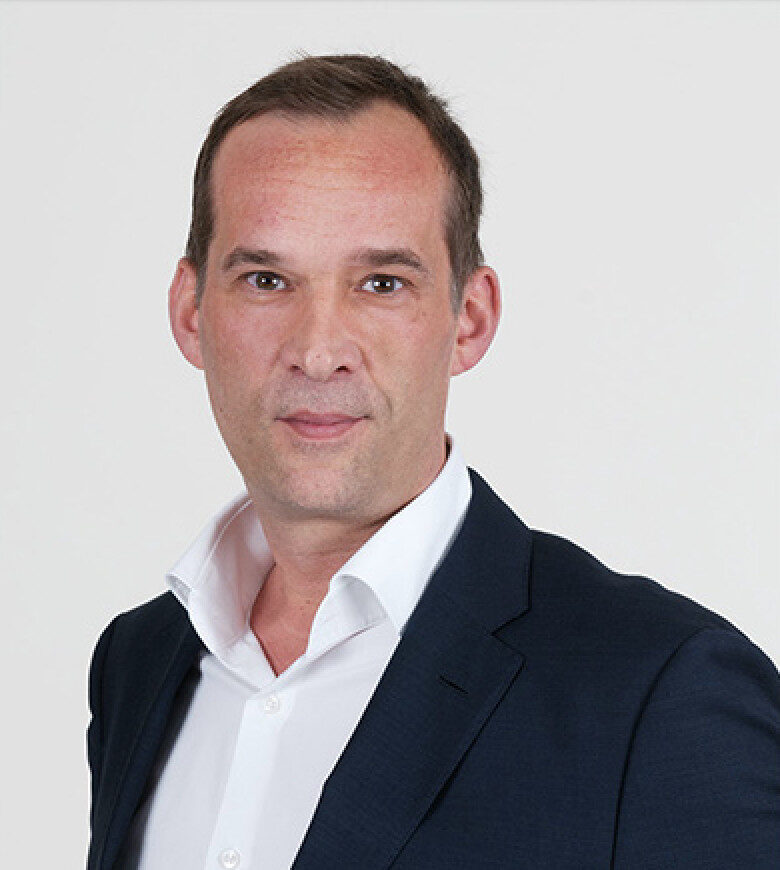 Portrait of a man in a suit with an open collar in front of a neutral background.