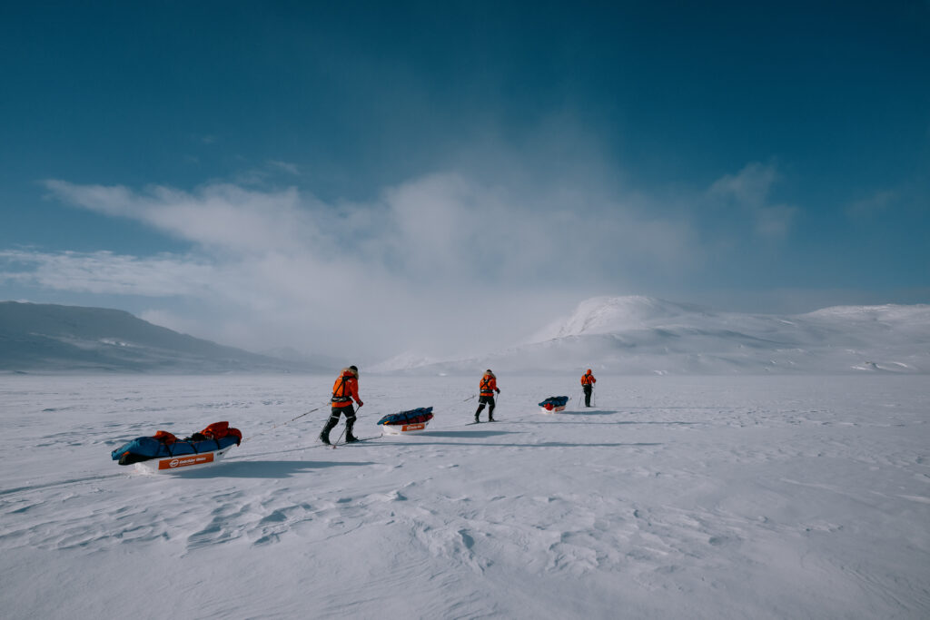 Vier Personen in orangefarbener Kleidung ziehen Schlitten über eine verschneite Landschaft.