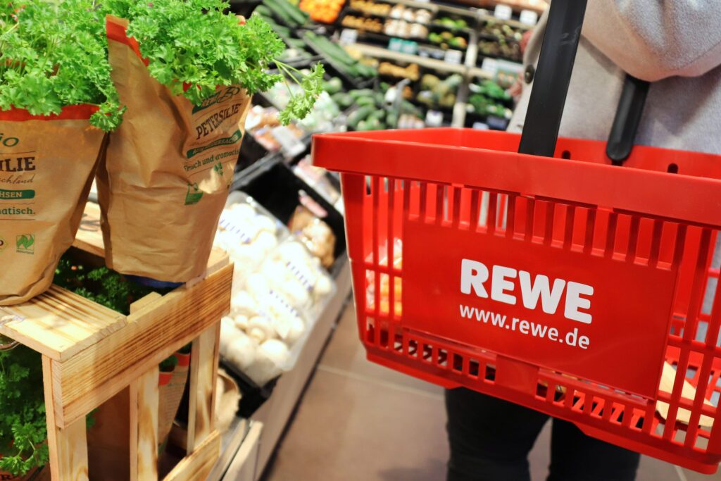 Roter Einkaufswagen von REWE mit frischen Kräutern und Gemüse im Hintergrund.