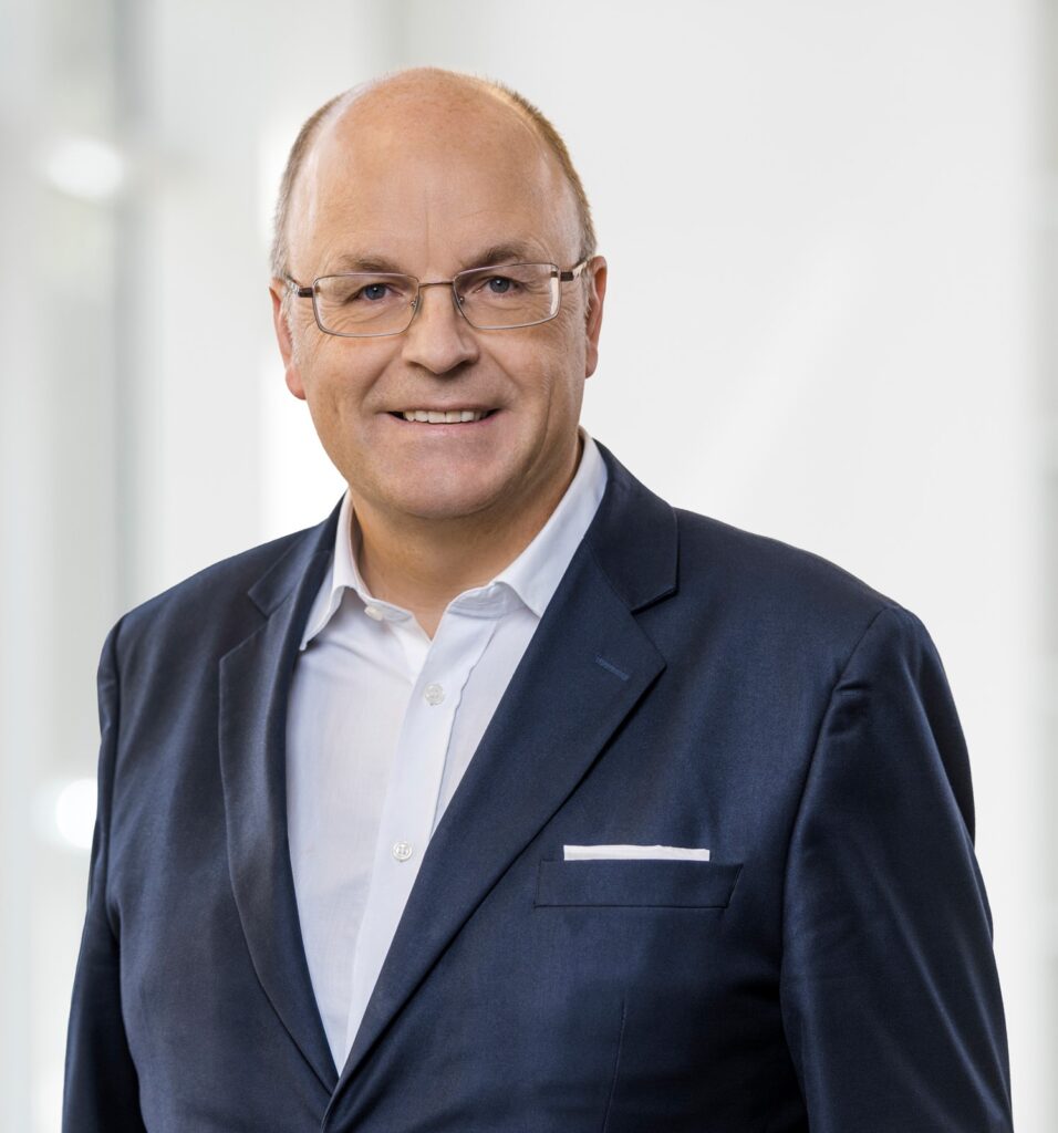 Porträt von S. Hofstetter in einem blauen Anzug mit Brille und lächelndem Gesicht.