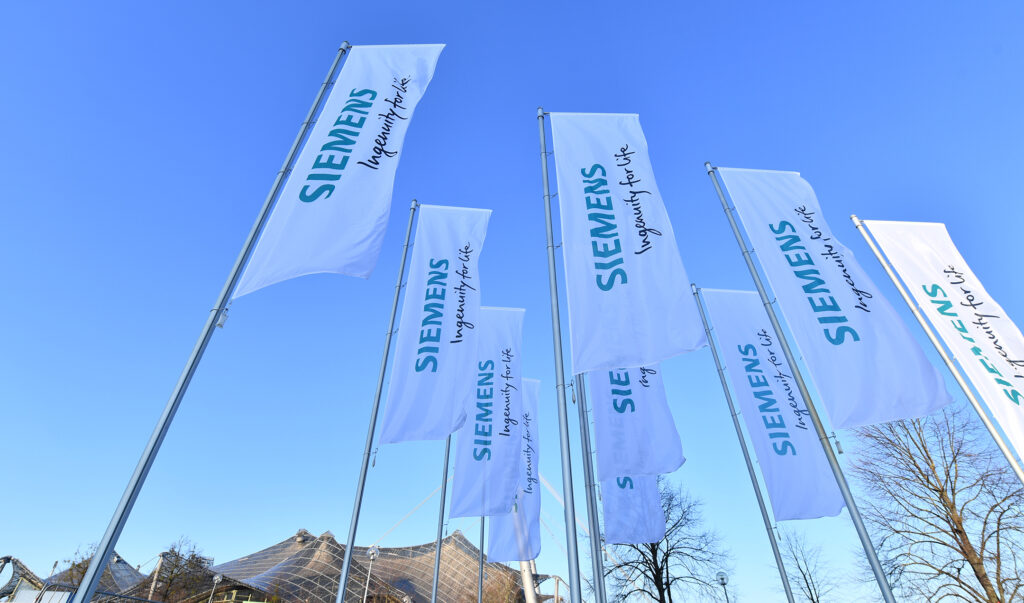 Mehrere Fahnen mit dem Logo von Siemens wehen im Freien unter blauem Himmel.
