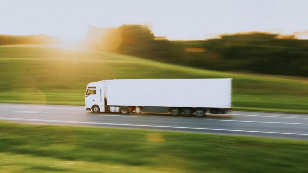 Ein weißer Lkw fährt auf einer Straße mit verschwommenem Hintergrund und Sonnenlicht.