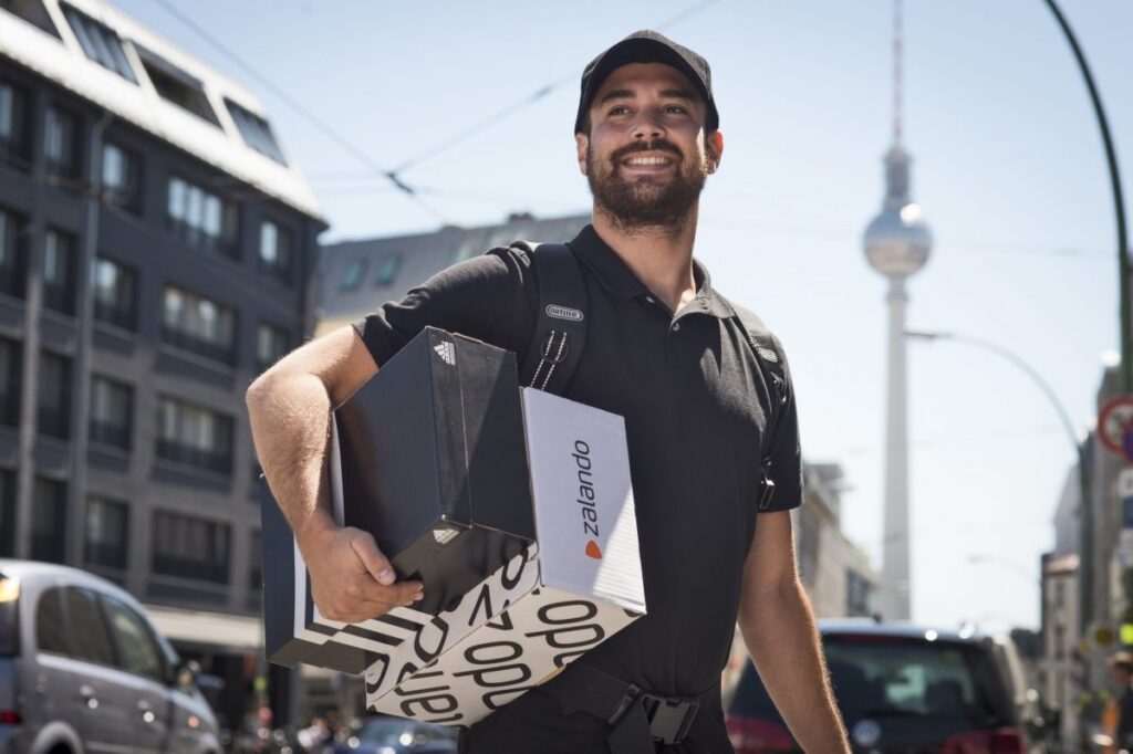 Kurier mit Zalando und Adidas Paketen in der Hand auf der Straße.
