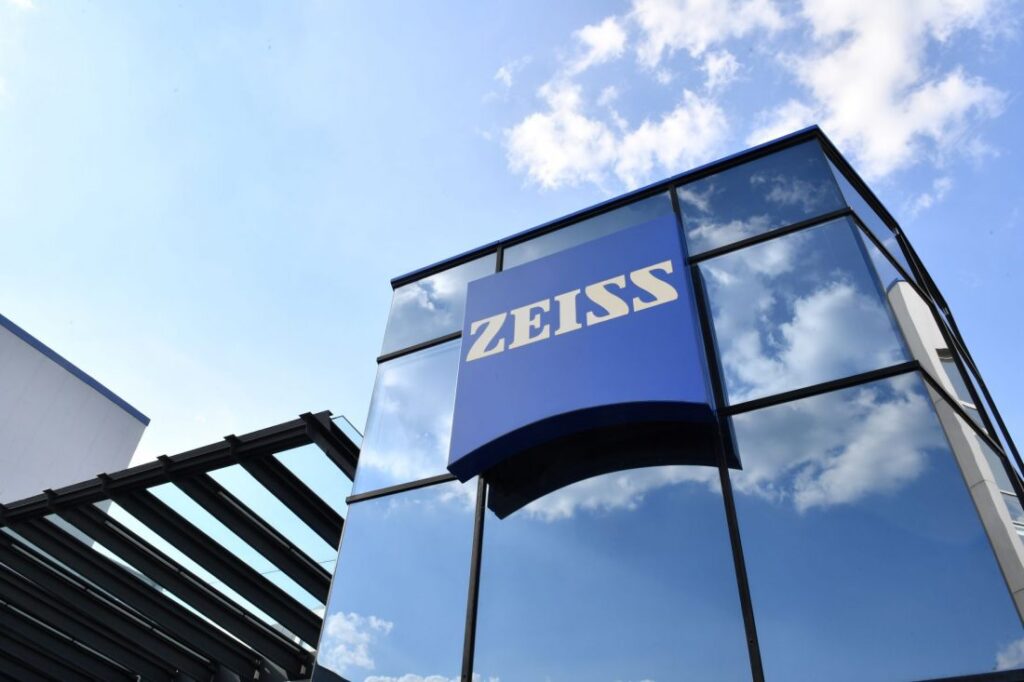 Gebäude mit dem Logo von Zeiss auf einer reflektierenden Glasfassade unter blauem Himmel.