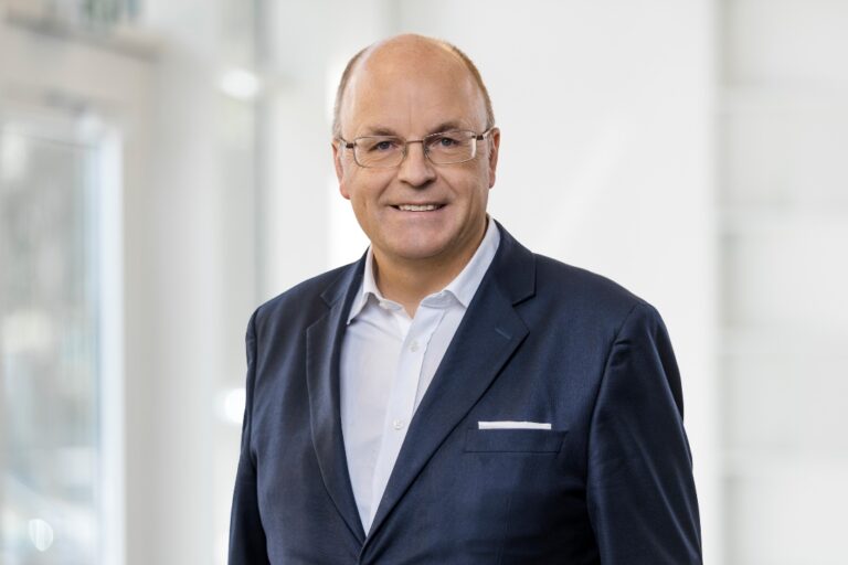 Mann in einem dunkelblauen Anzug mit Brille und lächelndem Gesicht vor neutralem Hintergrund.