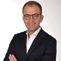Mann in einem schwarzen Anzug und Brille, der lächelt und die Arme verschränkt.