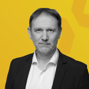 Porträt eines Mannes mit grauen Haaren und Bart, der einen schwarzen Anzug trägt.