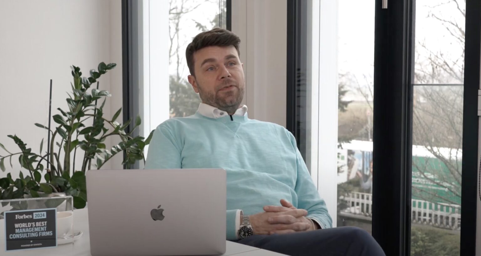 Mann in einem hellblauen Pullover sitzt an einem Tisch mit einem Laptop und spricht.