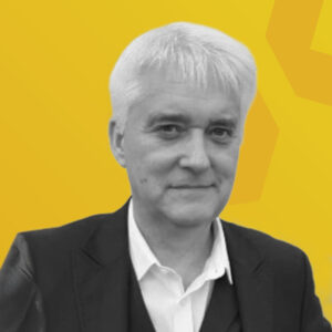 Porträt eines Mannes mit grauen Haaren, der einen schwarzen Anzug trägt, vor einem gelben Hintergrund.