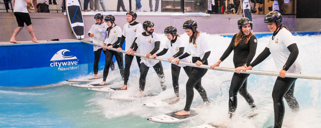 Eine Gruppe von acht Surfern steht auf Surfbrettern in der Citywave in Düsseldorf und hält sich an einer Stange fest.