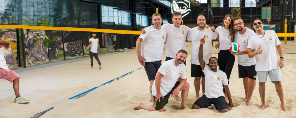 Eine Gruppe von Personen in weißen T-Shirts posiert auf einem Sandvolleyballfeld mit einem Volleyball.