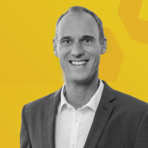 Mario Schmidtgen en costume gris et chemise blanche sur fond jaune.