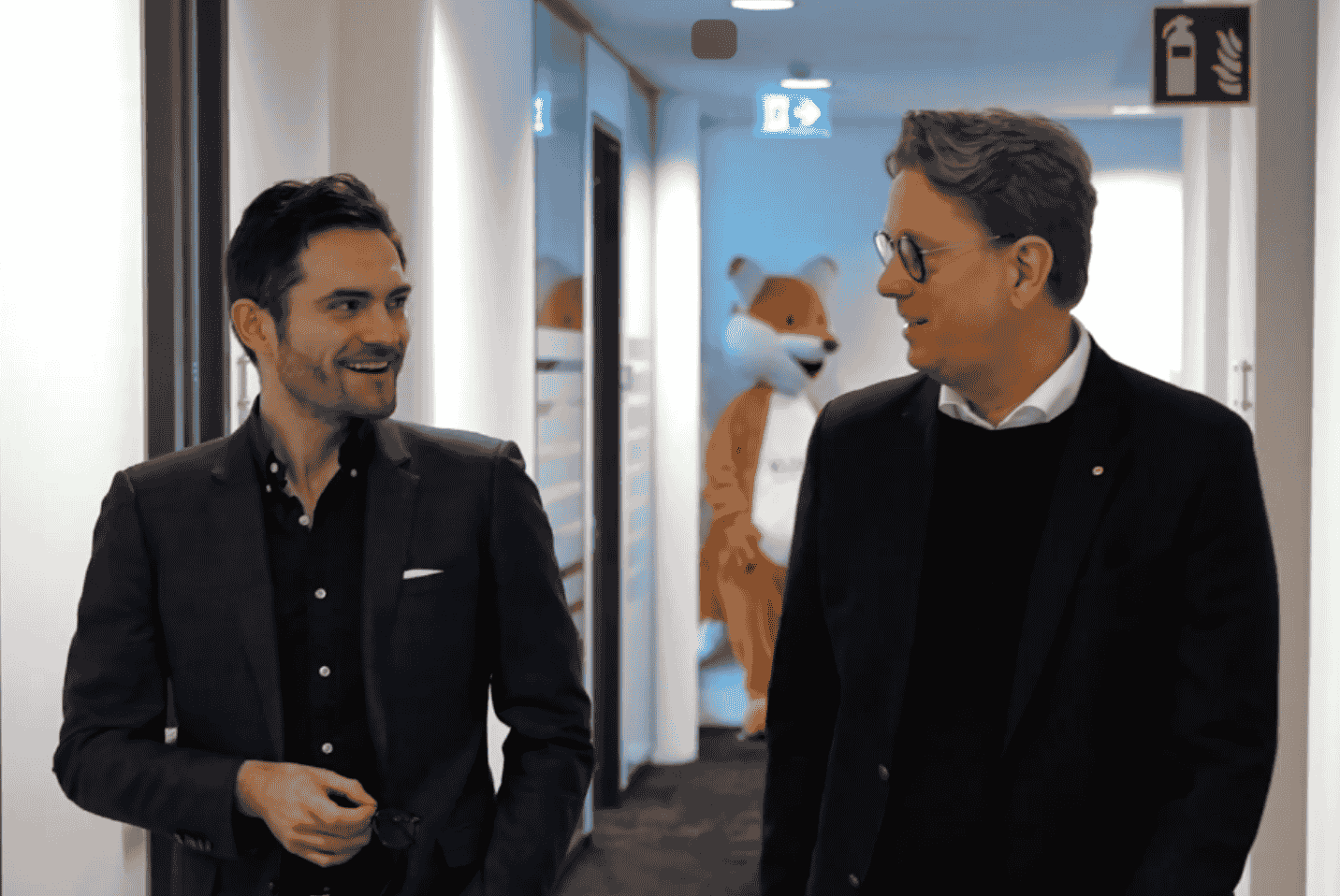 Deux hommes en costume qui marchent dans un couloir de bureau et discutent entre eux.