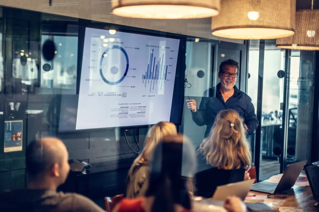 Ein Mann präsentiert Daten auf einem Bildschirm in einem Besprechungsraum mit mehreren Zuhörern.